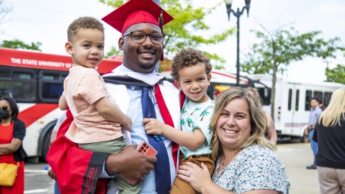 Graduate smiles as he looks on towards his two small children in his arms and spouse by his side