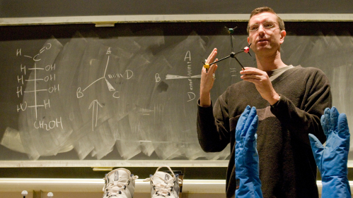 Professor stands in front of board showing CH2OH and holding model of molecutle with sneakers and gloves on table.
