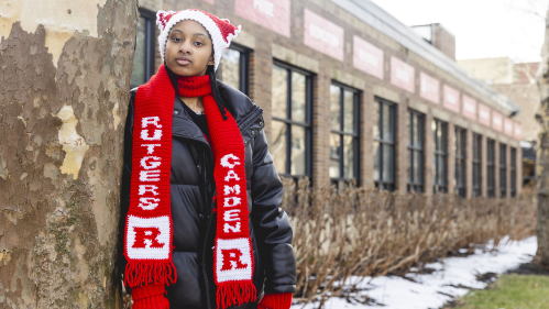 Student leans against tree by Campus Center wearing crocheted Rutgers hat and scarf.
