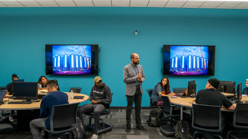 Students turn to look at professor in front of teal wall with 2 large tv monitors displaying same vibrant images.