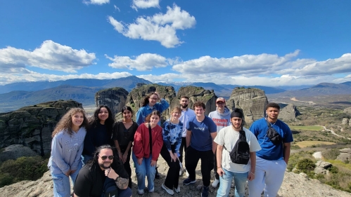 Rutgers-Camden Greece spring break field trip. Pictured in front of mountains with blue sky and while clouds.