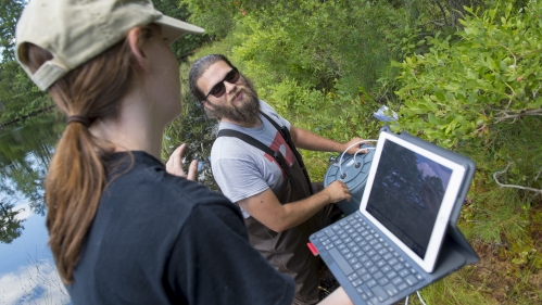 one male college student working on a container while a female college student documents on her tablet screen on the edge of a river