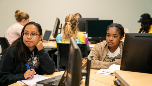 Students sit at round table with computers and notebooks.