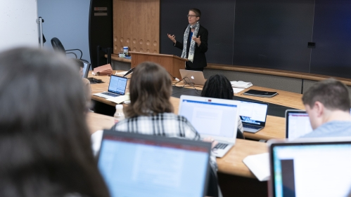 a female professor at the front of a lecture hall and the backs of college students taking notes in their open laptops