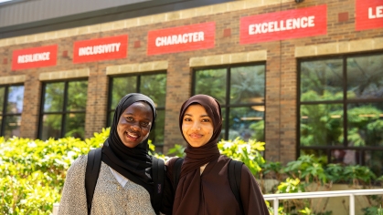 Two students with hijabs standing in front of the Campus Center. In the background are signs with the following words: Resilience, Inclusivity, Character and Excellence.