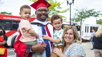 Graduate smiles as he looks on towards his two small children in his arms and spouse by his side
