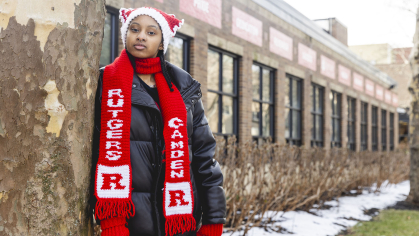 Student leans against tree by Campus Center wearing crocheted Rutgers hat and scarf.