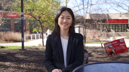 Professor wears a black blazer and sits outside on the campus quad