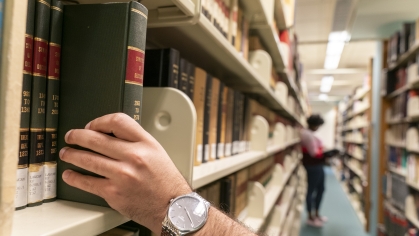 the hand of an adult male pulling a book off the law library shelves and an adult female reading a book in the background
