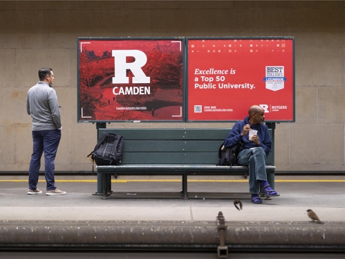 Passenger sitting on bench in front of Rutgers-Camden billboard display. Another standing passenger looks on to billboard as well.