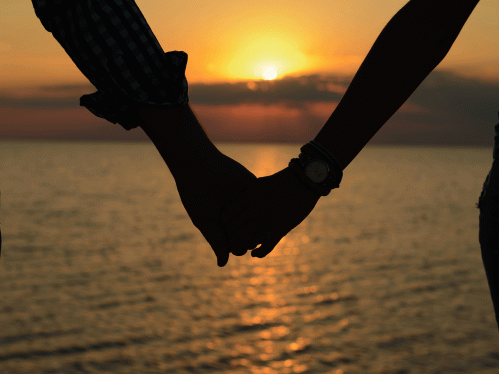 A loving couple (silhouettes of a man and a girl) hold hands in the rays of light at sunset on the sea on a pier.