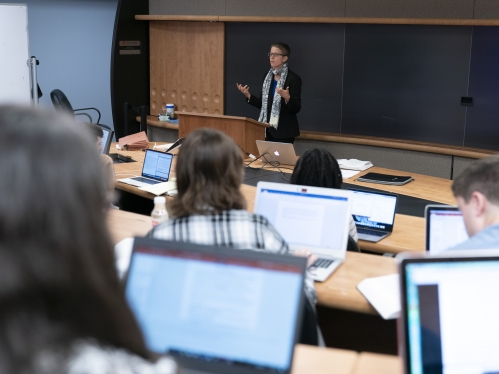 a female professor at the front of a lecture hall and the backs of college students taking notes in their open laptops