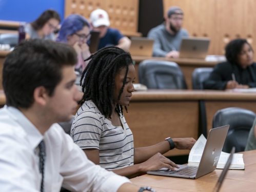 a female college student looking at her text book and taking notes on her laptop in a lecture hall with other college students