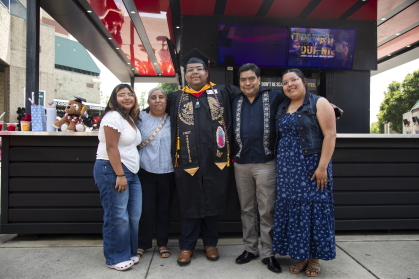 Male student in commencement regalia standing with his family with their arms around each other smiling with joy and pride. 