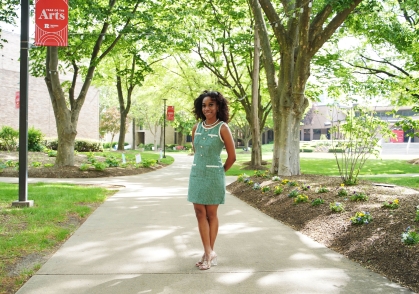 Student in green dress stands on a campus quad walkway.