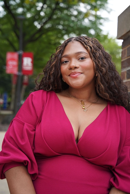 Woman wearing a red dress leans against a brick post with Rutgers banners in distance