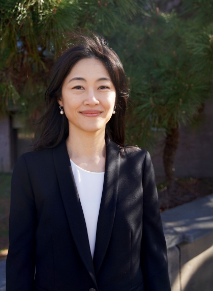 Professor wears a black blazer and is smiling in front of pine trees