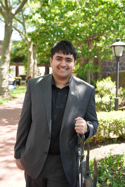 Shaan Mody wearing a gray suit leans against a fence by the Admissions building